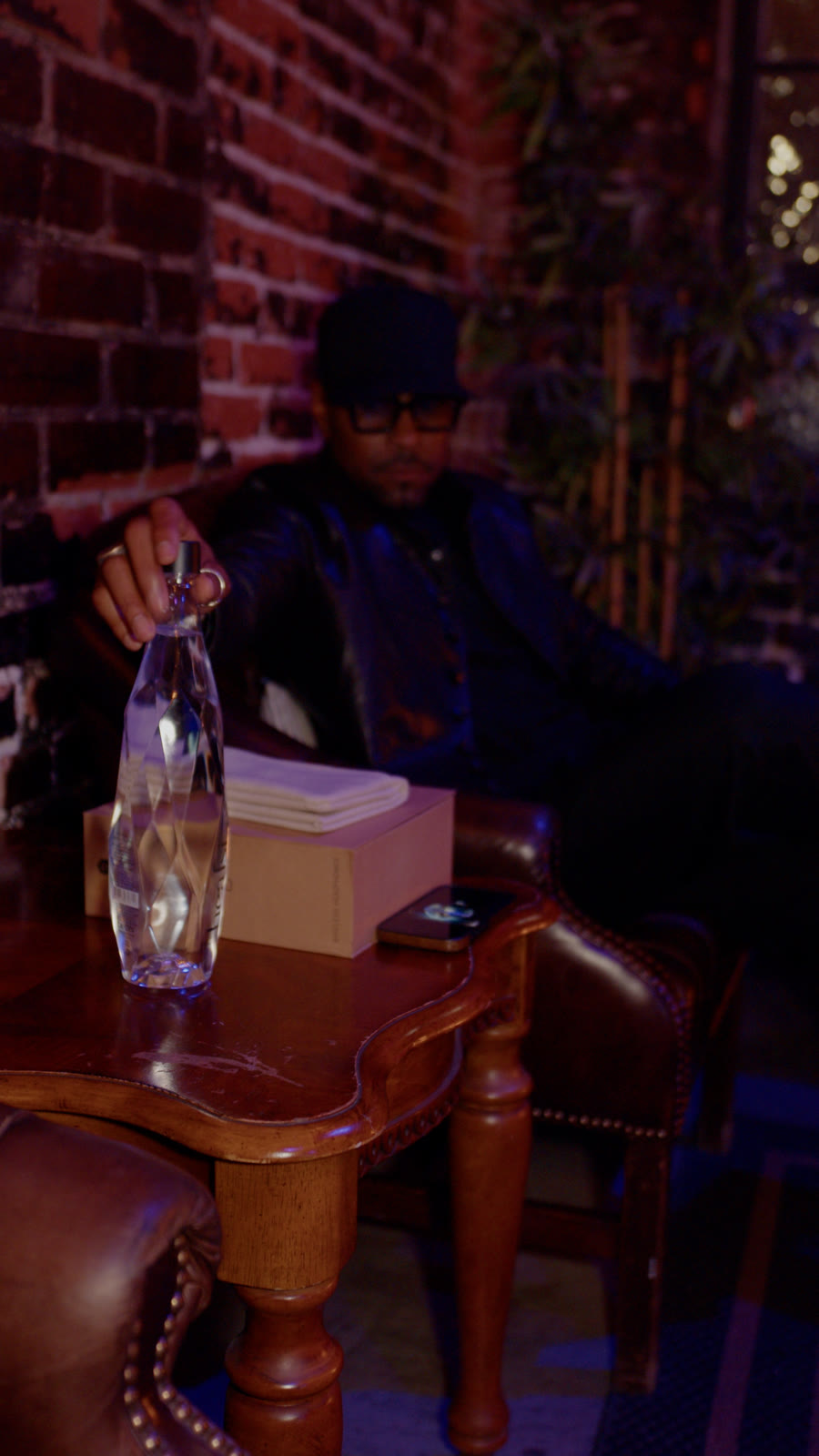 Aaron Skyy seated in a leather chair beside a side table in a dim brick lounge