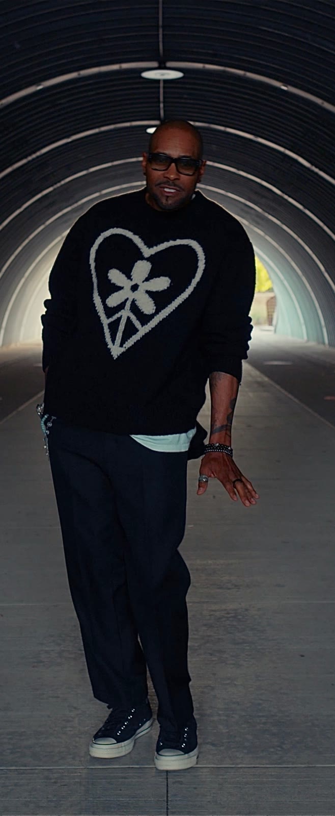 Aaron Skyy walking toward the camera through a corrugated tunnel in a heart-and-flower sweater
